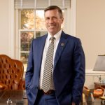 Dr. Hill smiling while standing in an office with a large wooden desk, brown leather chair, and various office items