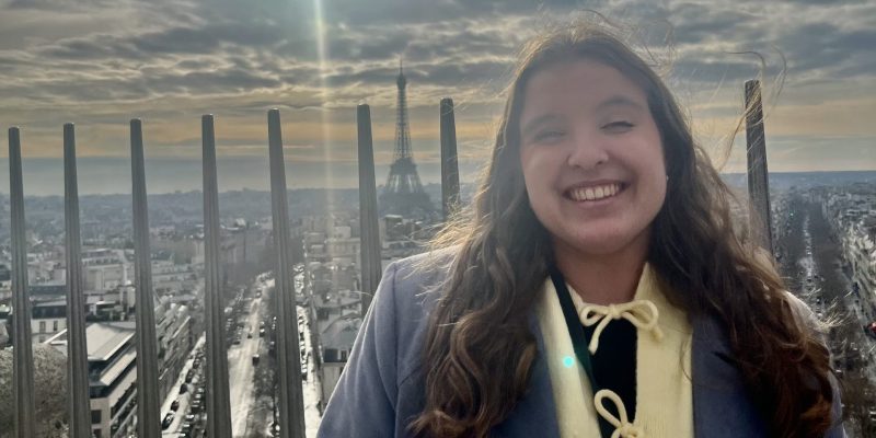 Person smiling in front of metal railings with the Eiffel Tower and a bright sun in a cloudy sky behind.