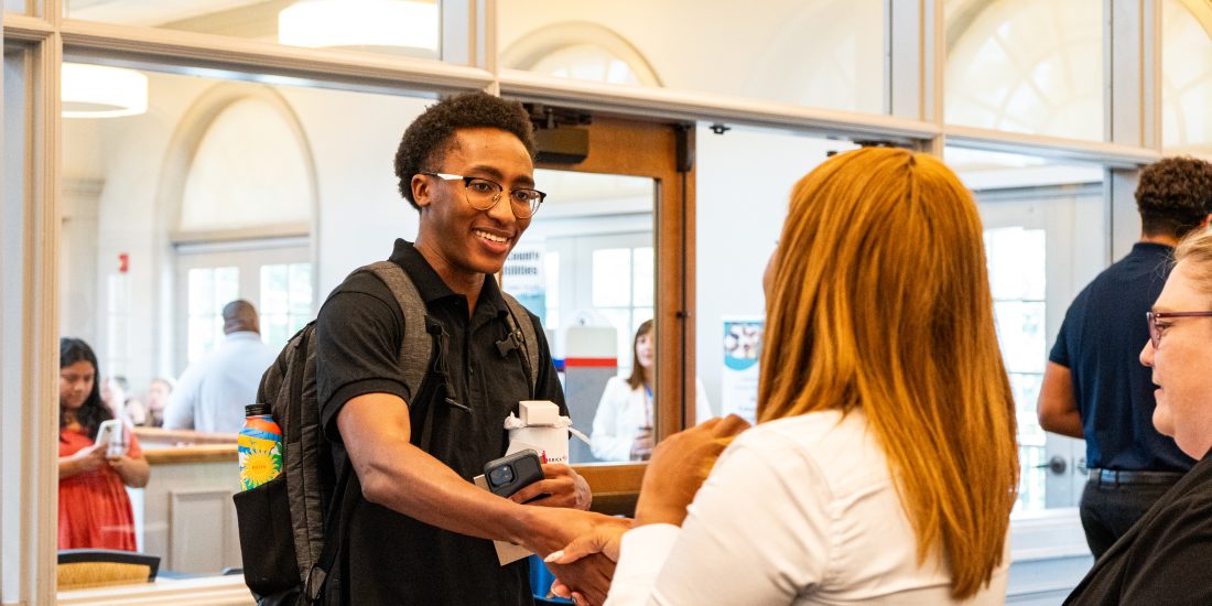 Two people shake hands inside a brightly lit building as groups of people stand and talk in the background.