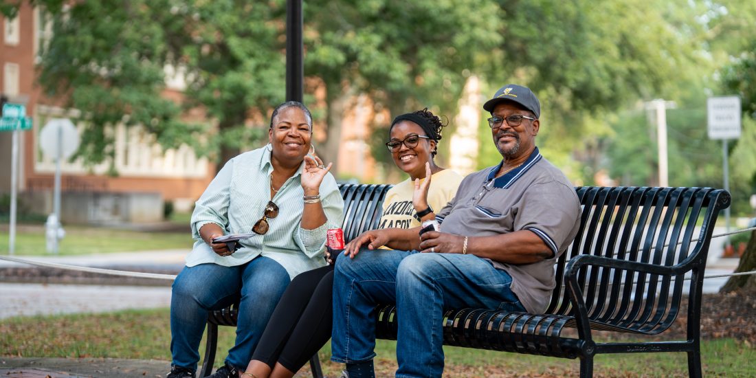 Three individuals sit and smile on a bench, smiling and waving at the camera. Trees and buildings are visible in the background.