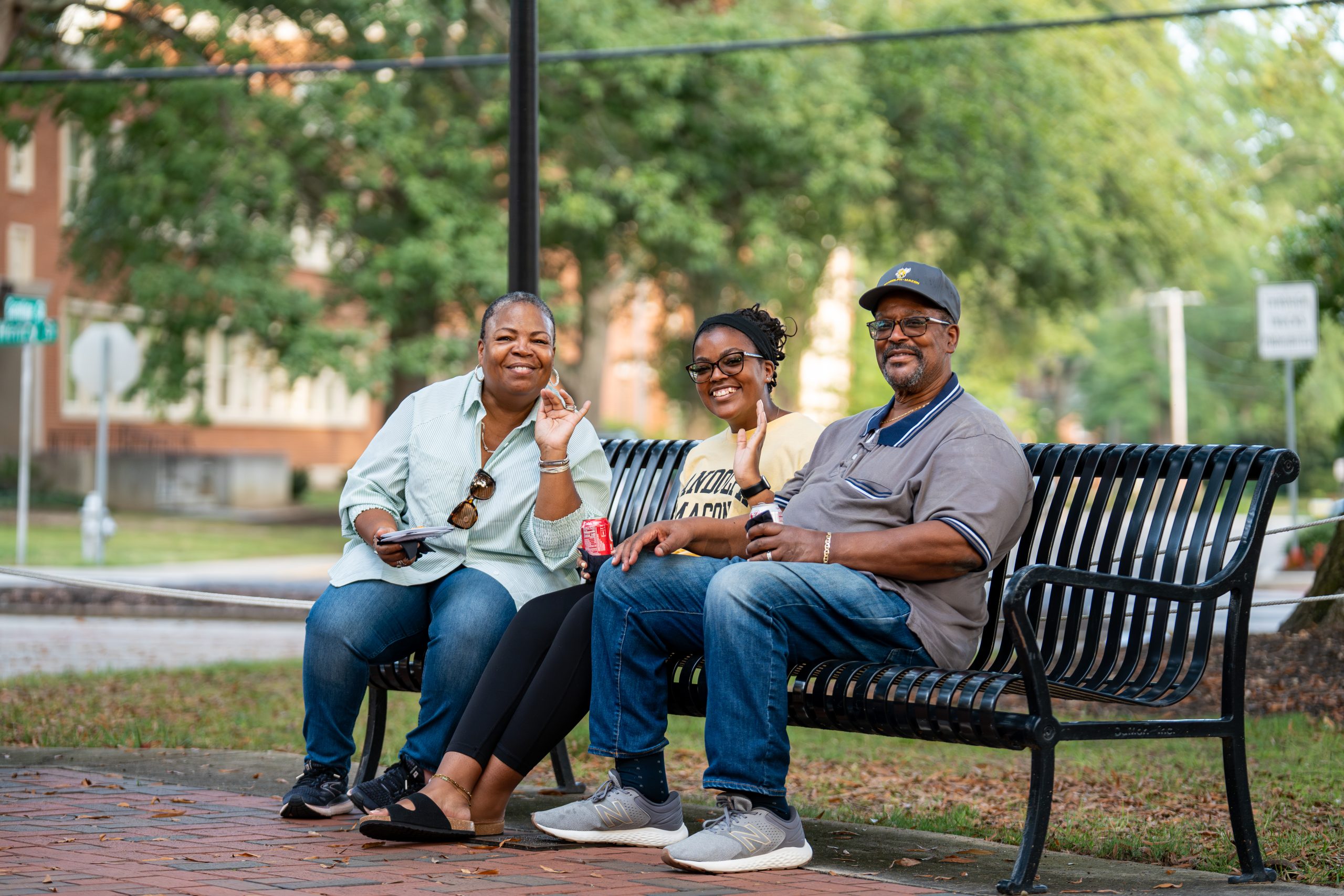 Three individuals sit and smile on a bench, smiling and waving at the camera. Trees and buildings are visible in the background.