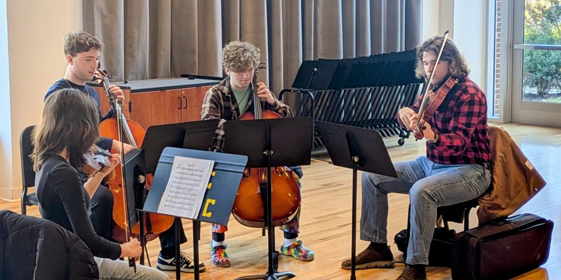 Musicians rehearse together in a well-lit room, playing two cellos, a viola, and a violin with sheet music on stands.