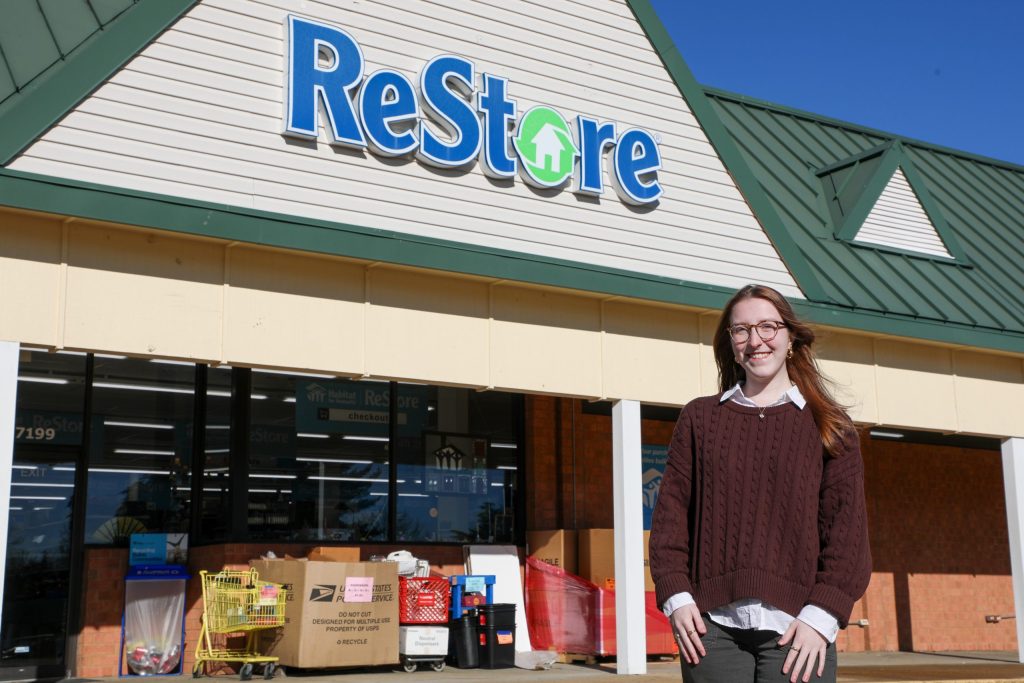 A young woman stands outside a ReStore retail building with shopping carts and donation items near the entrance.