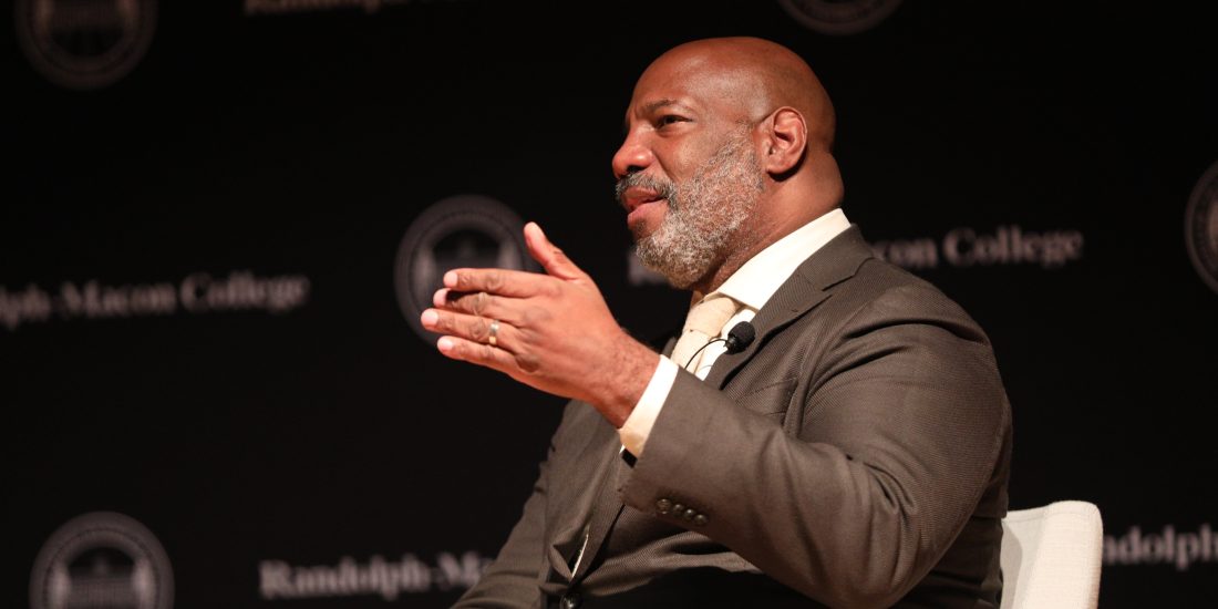 A person speaks on stage at an event, gesturing, with a backdrop displaying the words "Randolph-Macon College.