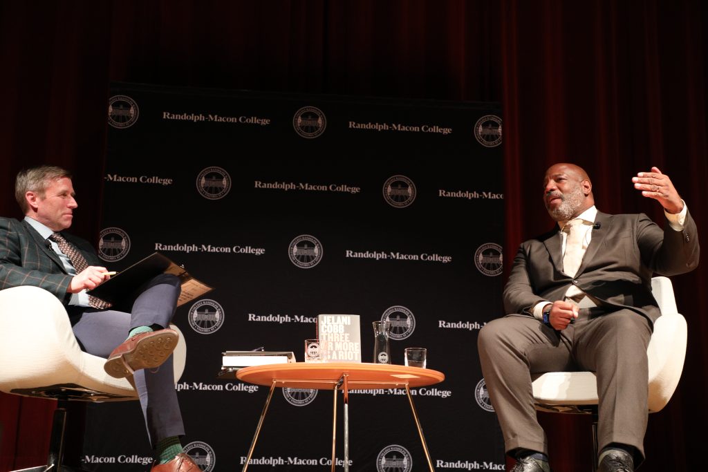 Two people have a discussion on stage at Randolph-Macon College, with the book "Three Or More Is A Riot" displayed on the table between them.