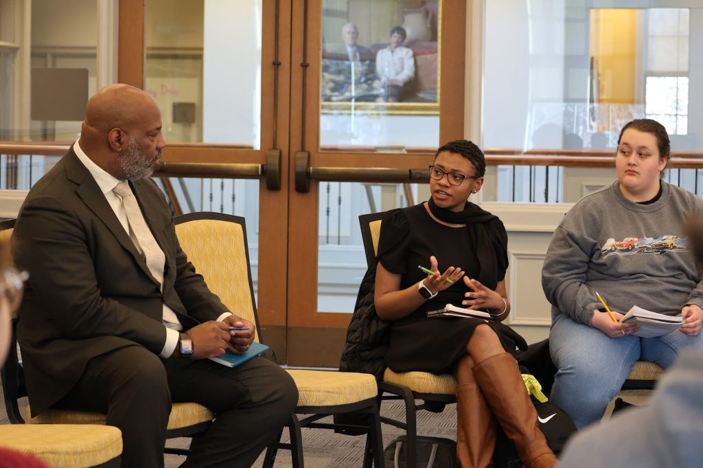 Three people sit indoors in a discussion circle; one speaks with hand gestures, another holds a notebook and pen.