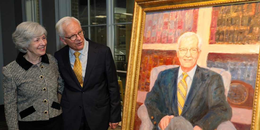 Two people stand beside a framed painted portrait, displayed indoors
