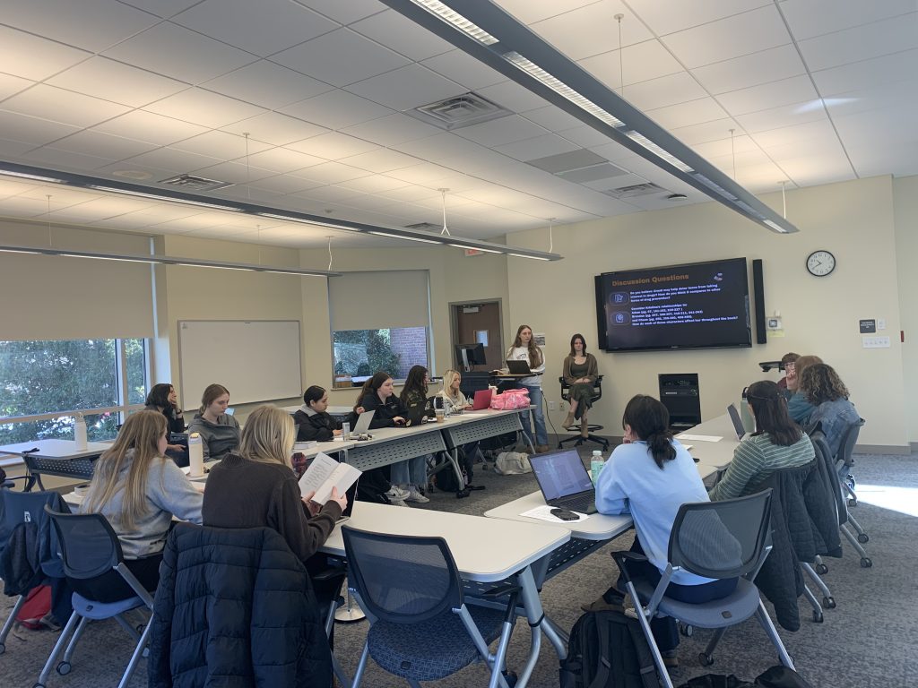A classroom with students at tables working or reading, and two individuals presenting near a screen showing discussion questions.