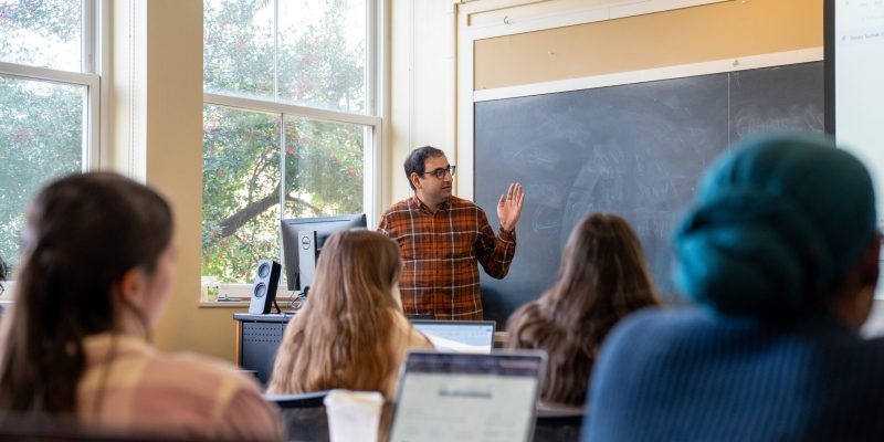An instructor addresses students seated at desks with open laptops in a classroom setting.