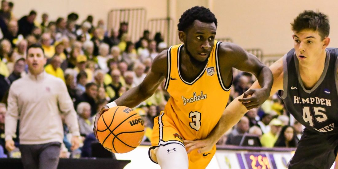 A Randolph-Macon basketball player dribbles past a Hampden-Sydney defender inside Crenshaw Gymnasium with crowded stands in the background.