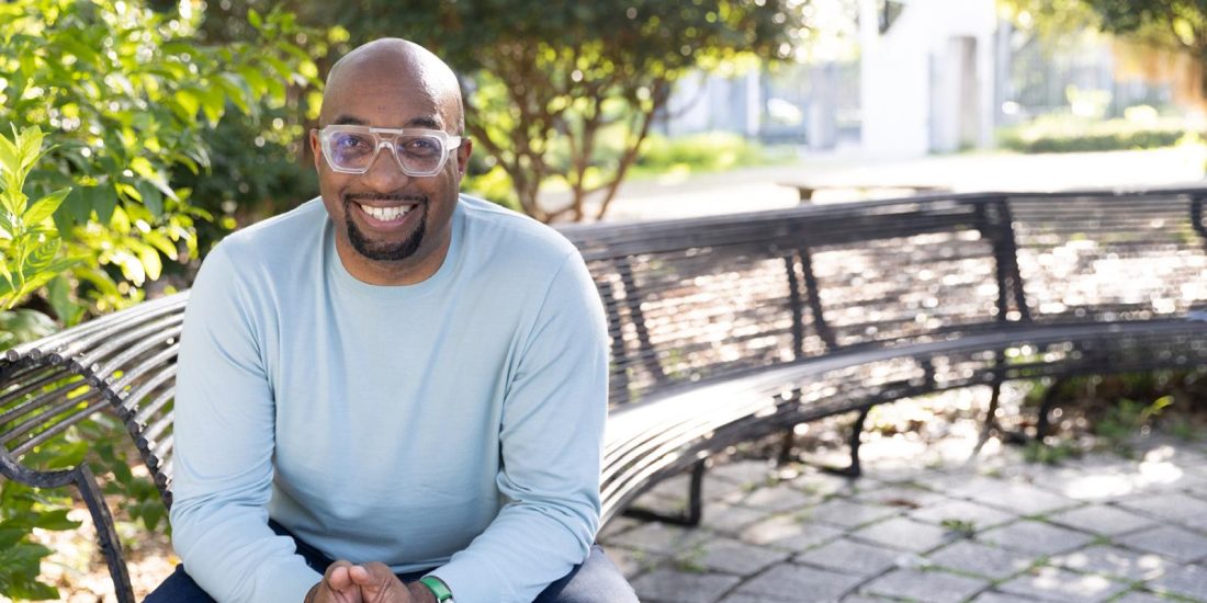 Person sits on a curved metal bench in a sunlit park, smiling at the camera.