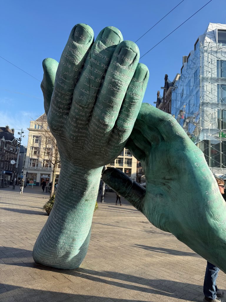 Bronze sculpture of two upright hands with engraved text, displayed in a city square amid buildings and glass structures.