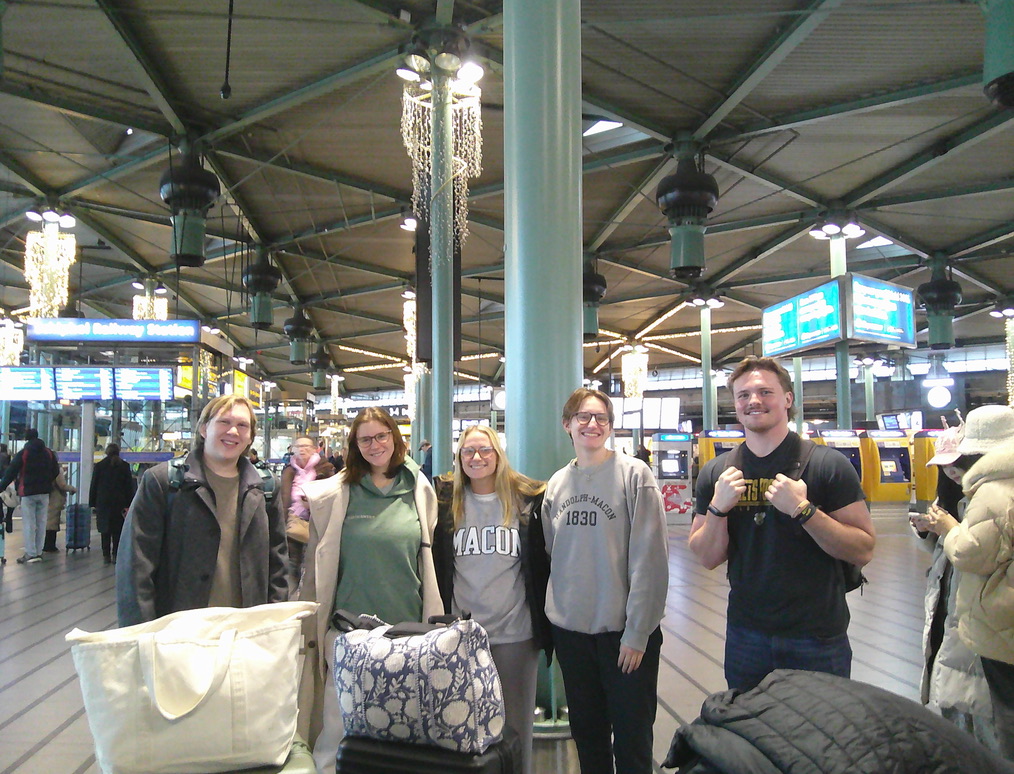Five people with luggage stand together in a train station, facing the camera; electronic boards and travelers are in the background.
