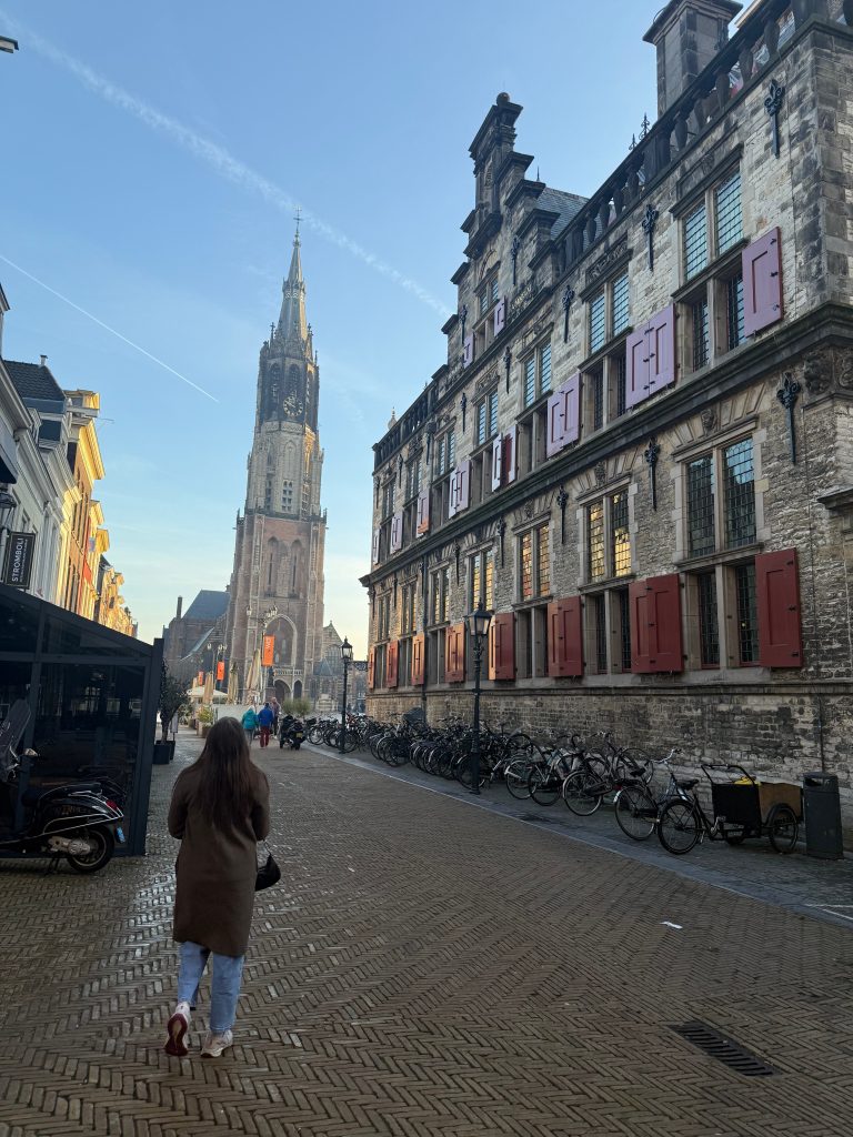 An individual walks along a cobblestone street with bicycles, historic buildings, and a church tower visible under a clear sky.