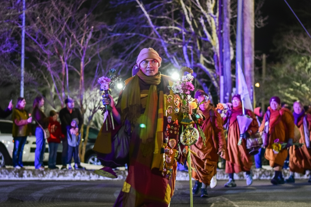 A monk walks at night holding flowers and lights, accompanied by others under streetlights.