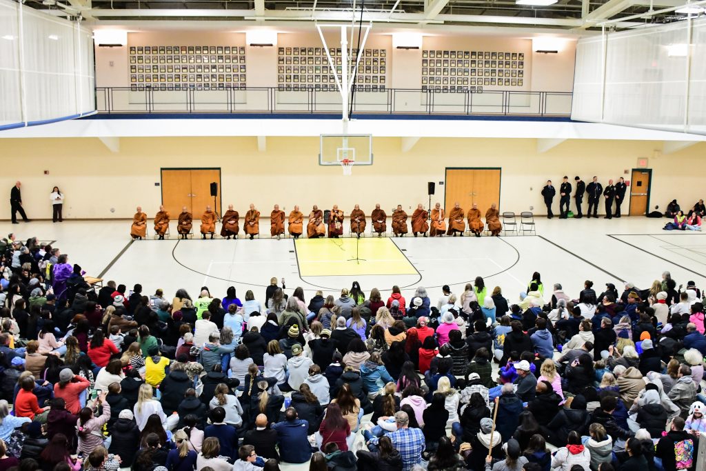 A crowd sits on a gymnasium floor, listening to monks seated in a row