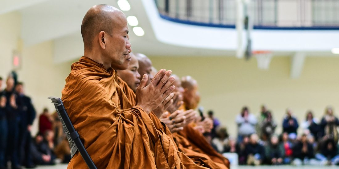 Buddhist monks sit with hands in prayer during an indoor ceremony, as an audience observes in the background.