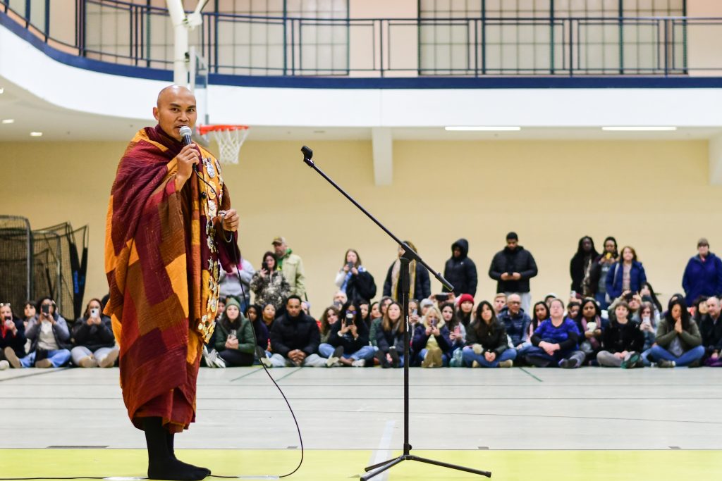 A monk speaks into a microphone on a gymnasium floor with an audience seated in the background.