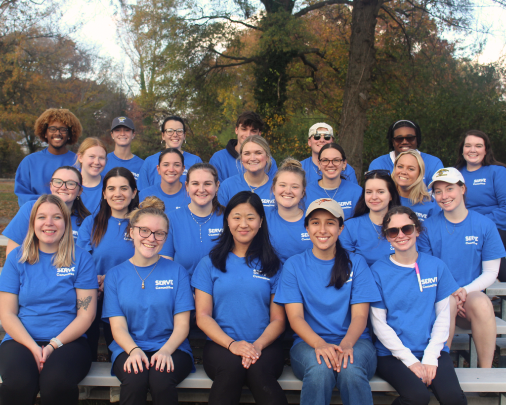 A group of twenty-two people in matching SERVE T-shirts pose outdoors on bleachers with autumn trees in the background.
