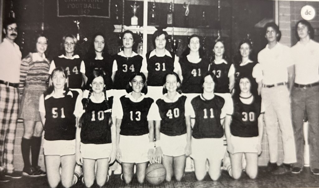 A black-and-white team photo of the 1975-76 RMC Women's Basketball team in front of a trophy case.