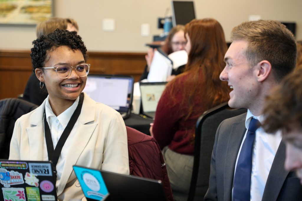Students in professional dress are seated around a circular table and smiling with laptops open in front of them
