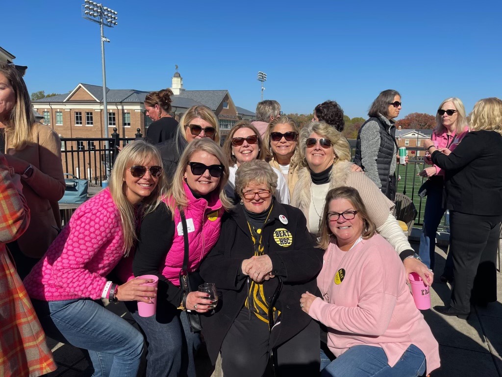 A group of smiling women wearing pink surround a seated woman in Randolph-Macon gear and a "Beat HSC" pin