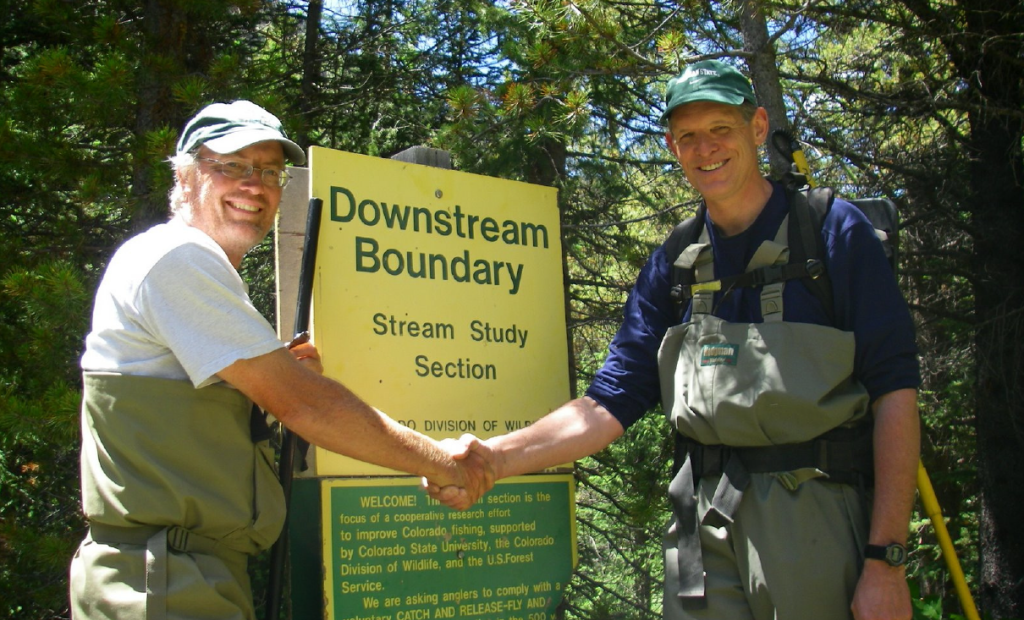 Dr. Chas. Gowan and Dr. Kurt Fausch shake hands in front of a yellow sign that reads "Downstream Boundary"