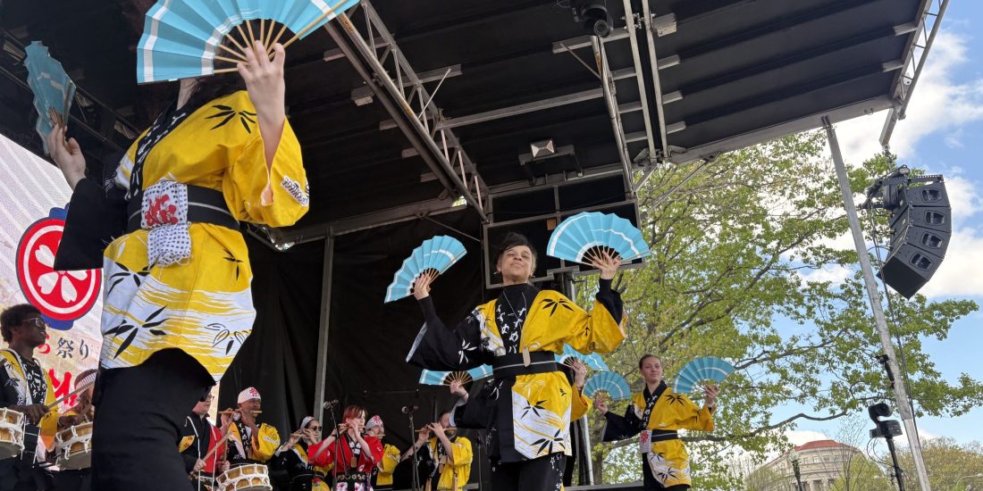 Students wearing traditional Japanese clothing, colored yellow and black, dance with fans on a stage in Washington, D.C., with more students playing flutes and drums in the background