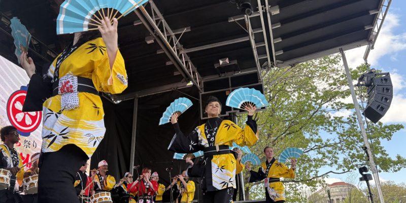 Students wearing traditional Japanese clothing, colored yellow and black, dance with fans on a stage in Washington, D.C., with more students playing flutes and drums in the background