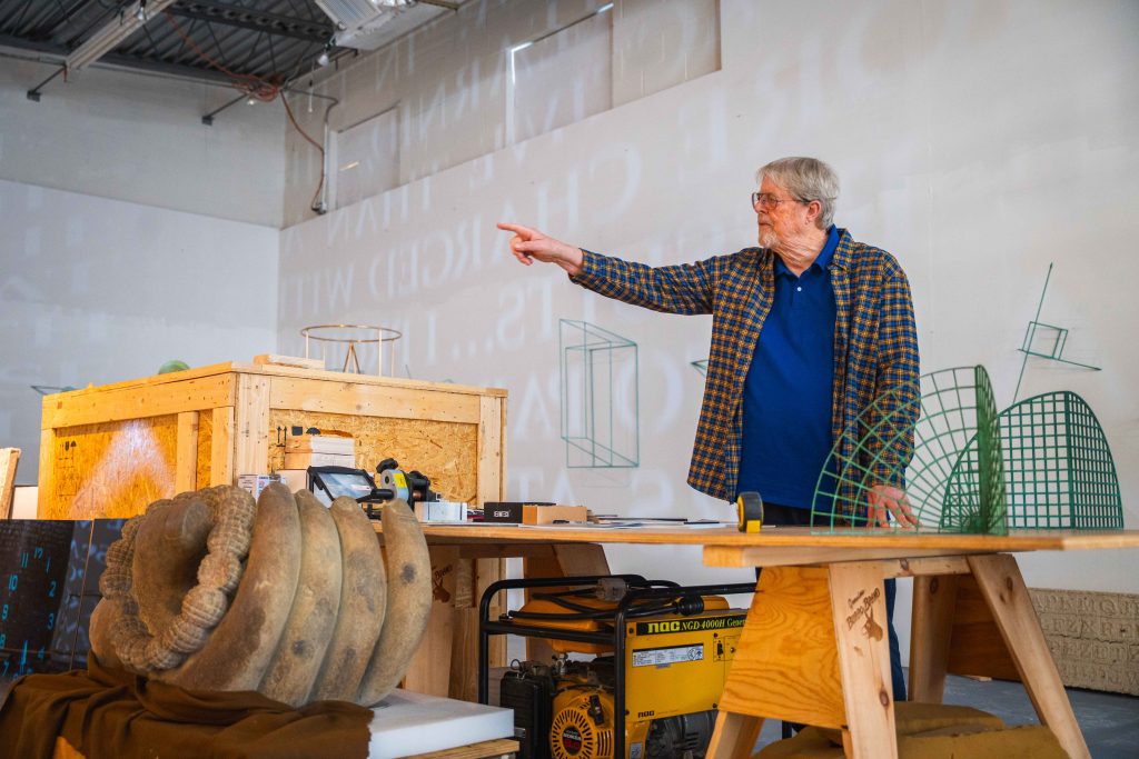 Surrounded by tools and sculptures in his studio, Jim Sanborn points towards the wall