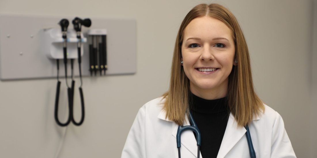 Madison Alderson stands smiling in an exam room wearing a white coat and a stethoscope around her neck.