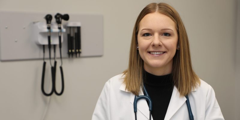 Madison Alderson stands smiling in an exam room wearing a white coat and a stethoscope around her neck.