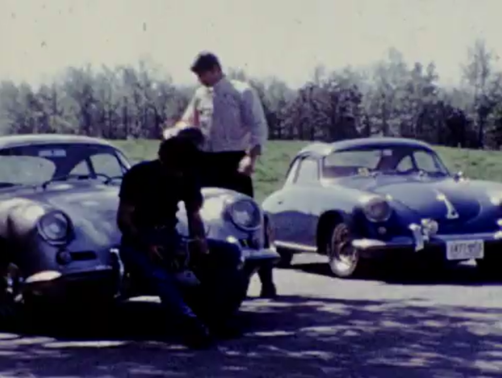 A screenshot of grainy footage of young men standing next to classic cars