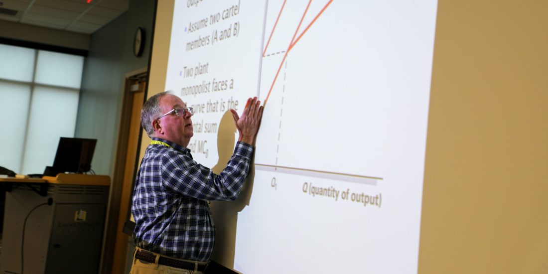 A professor stands in front of a classroom, demonstrating a curve on an economics graph