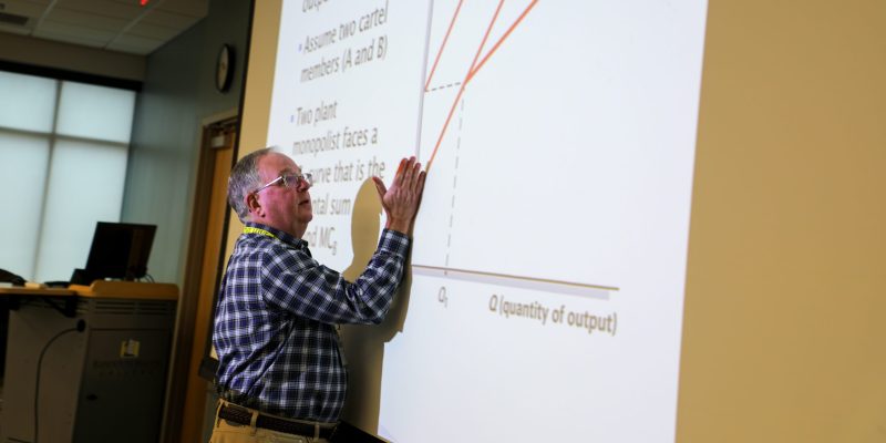 A professor stands in front of a classroom, demonstrating a curve on an economics graph