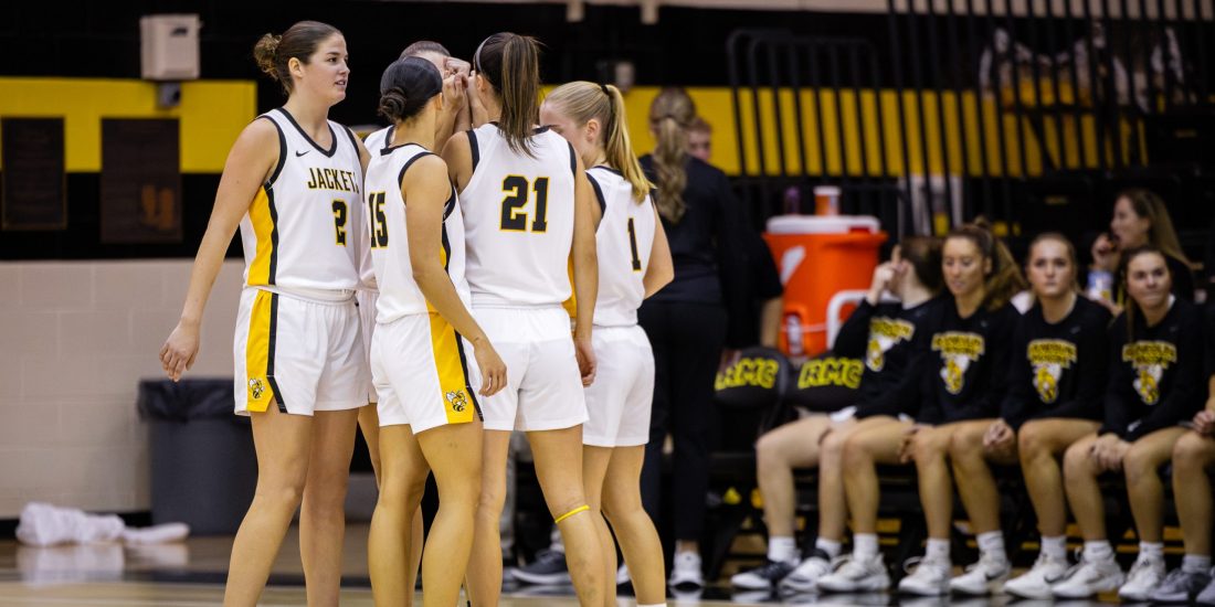 Five RMC Women's Basketball players huddle together on the court inside Crenshaw Gymnasium