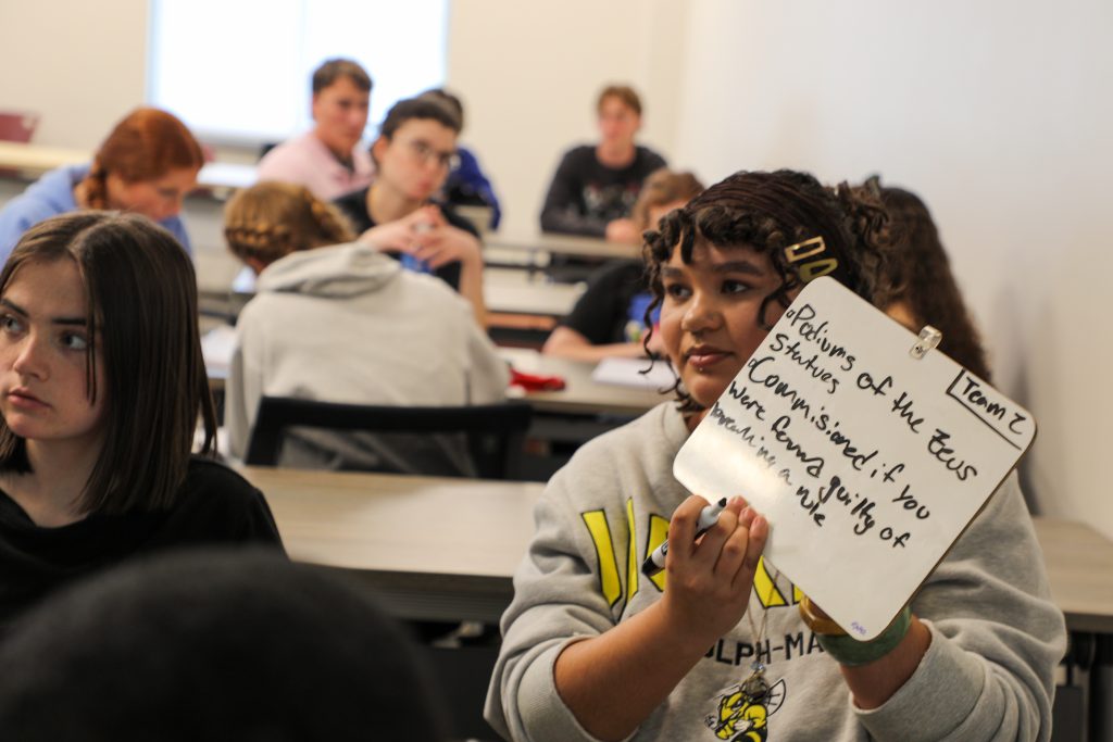 Students in groups hold up whiteboards with answers to training game questions in a Classics course
