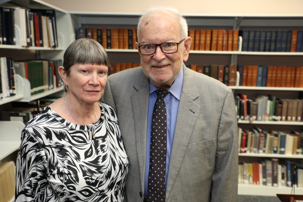 An elderly couple poses together inside a library with shelves of books in the background