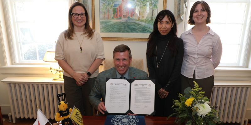 RMC President Michael Hill, flanked by College faculty and staff, holds a signed document while seating at a table displaying Japan and RMC flags, a Hirosaki University stole, and flowers