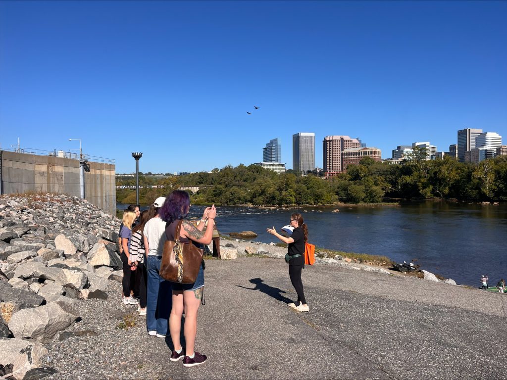 Patrons listen to a guided tour of the Manchester neighborhood with the James River and Richmond skyline in the background