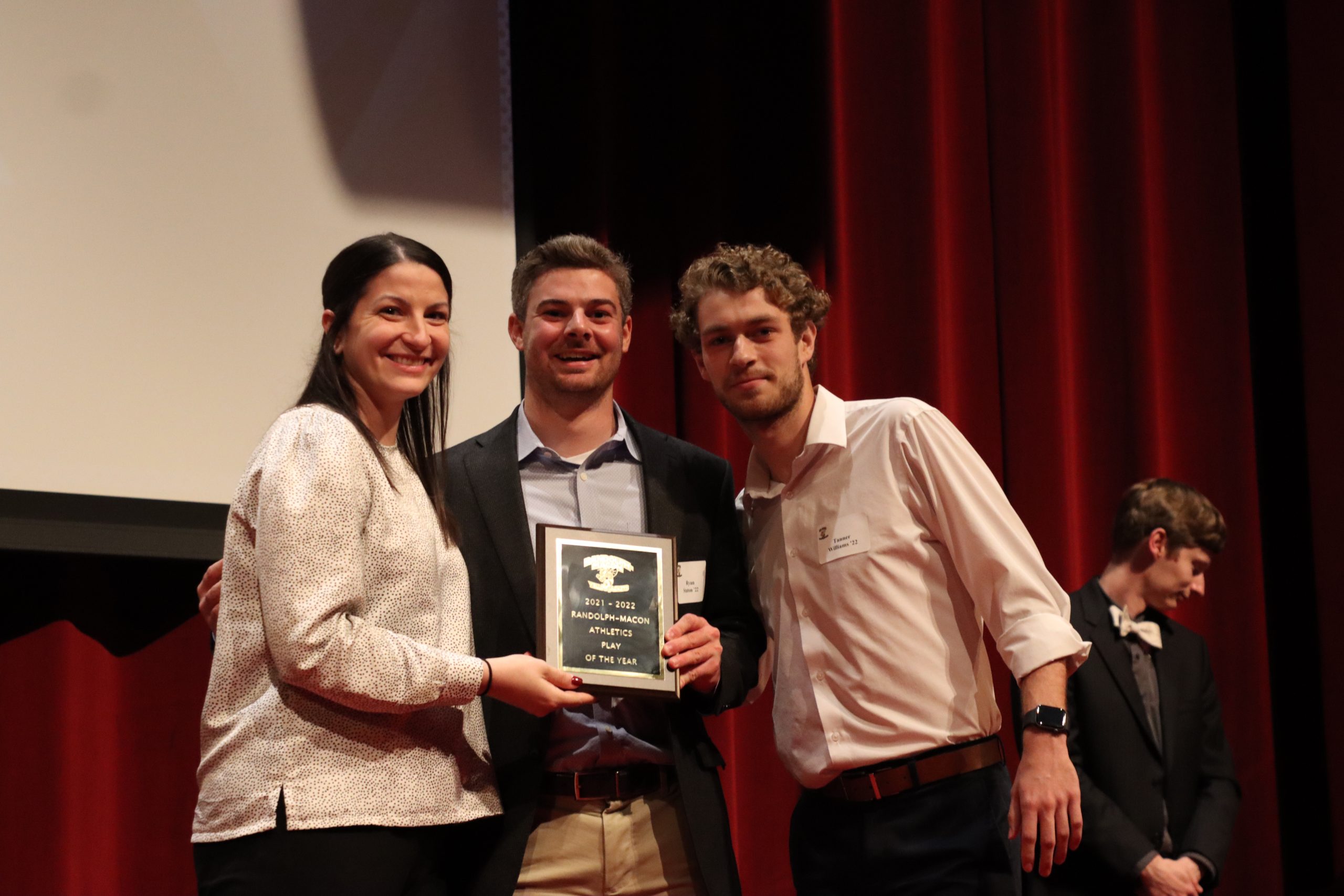 Three individuals stand on a stage during the Championship Award Ceremony holding an award plaque between them.