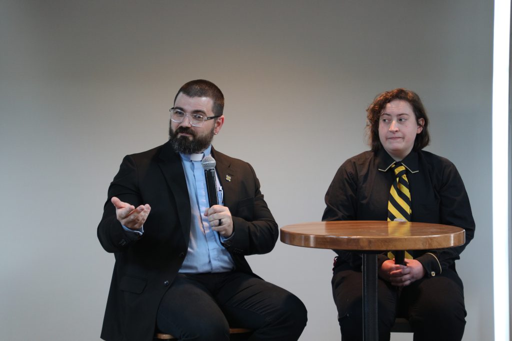A reverend talks into a microphone while seated at a high-top table next to a student