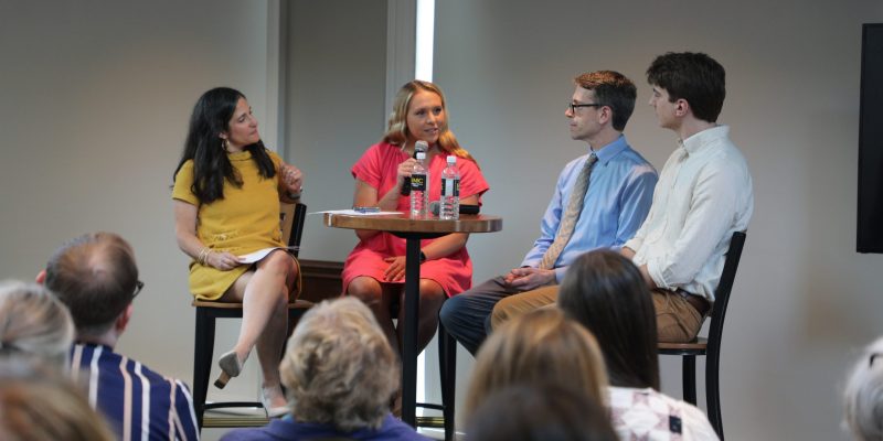 Four people sit at a high-top table having a discussion into microphones in front of a seated crowd