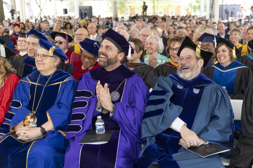 A seated crowd, with faculty members in regalia in the front rows, laughs and applauds