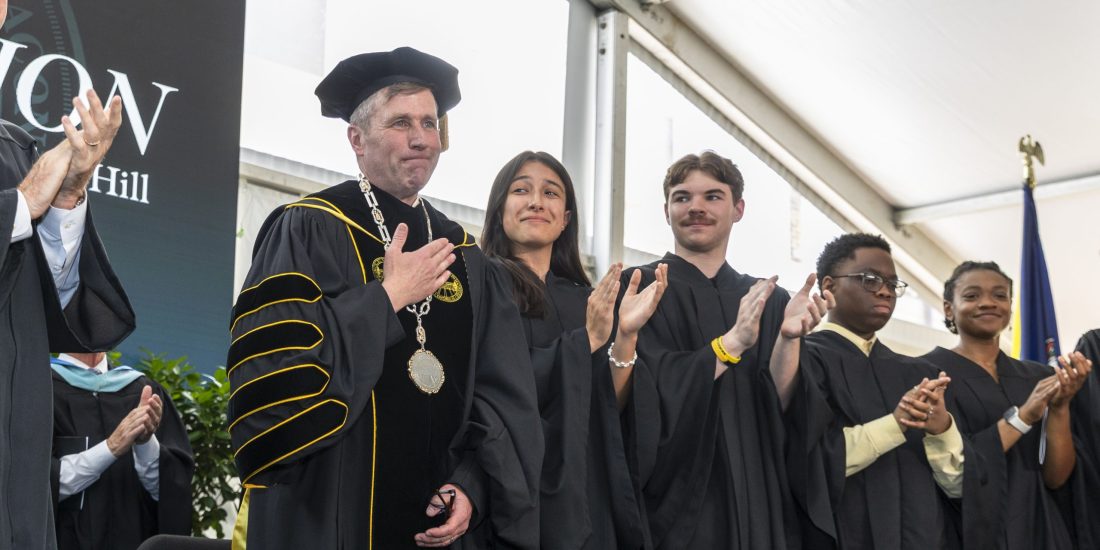 President Michael Hill, wearing the Presidential Medallion, puts a hand to his heart as students in regalia next to him applaud