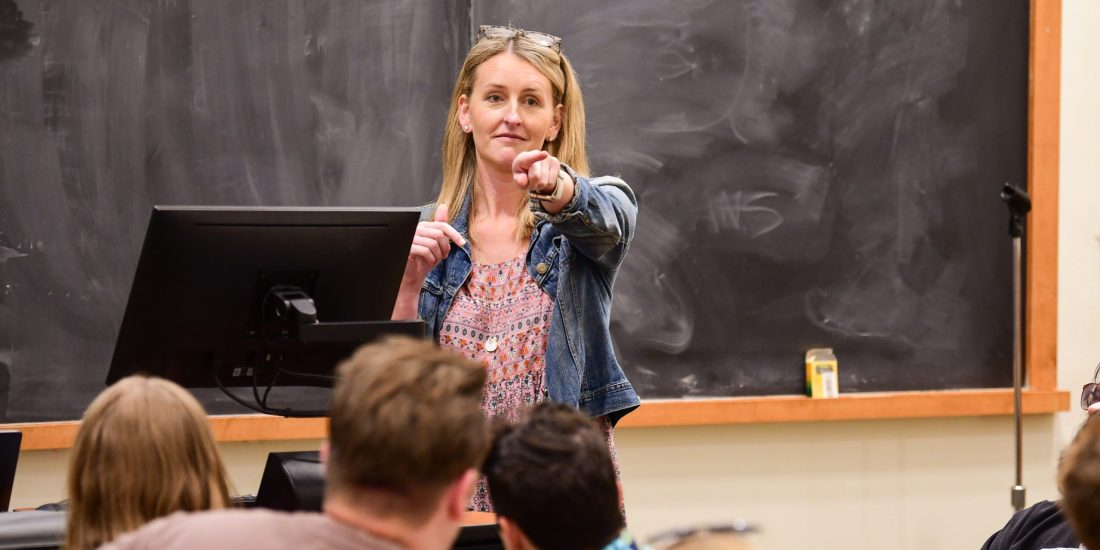 Maddie Smith stands in front of a blackboard and points to call on a student in a YJSS class