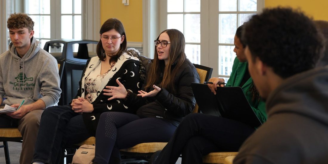 Students sit in circle of chairs having a discussion moderated by a professor
