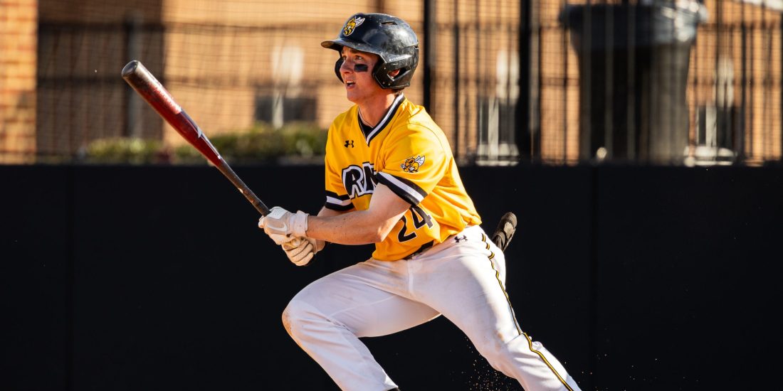 An RMC Baseball player holds a bat after a swing, running out of the batter's box