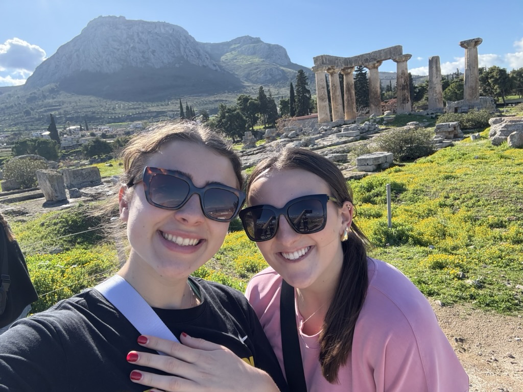 A selfie of two students standing in front of ancient Greek ruins on a sunny day