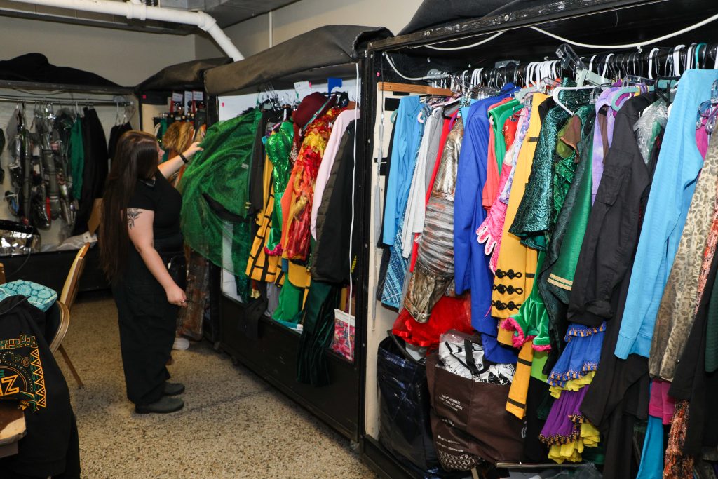 A young woman sorts through racks of colorful theater costumes in a backstage area